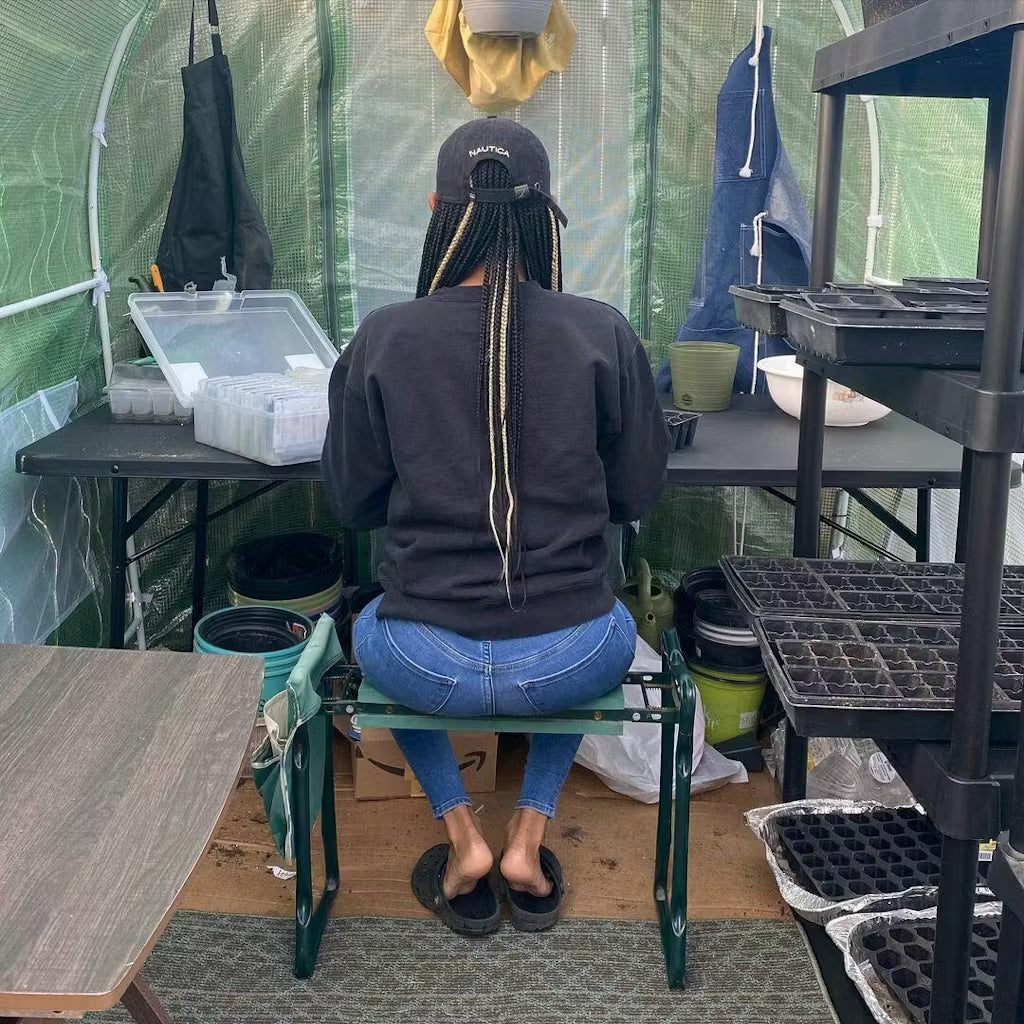 Person sitting on a stool in a greenhouse setting with gardening supplies around