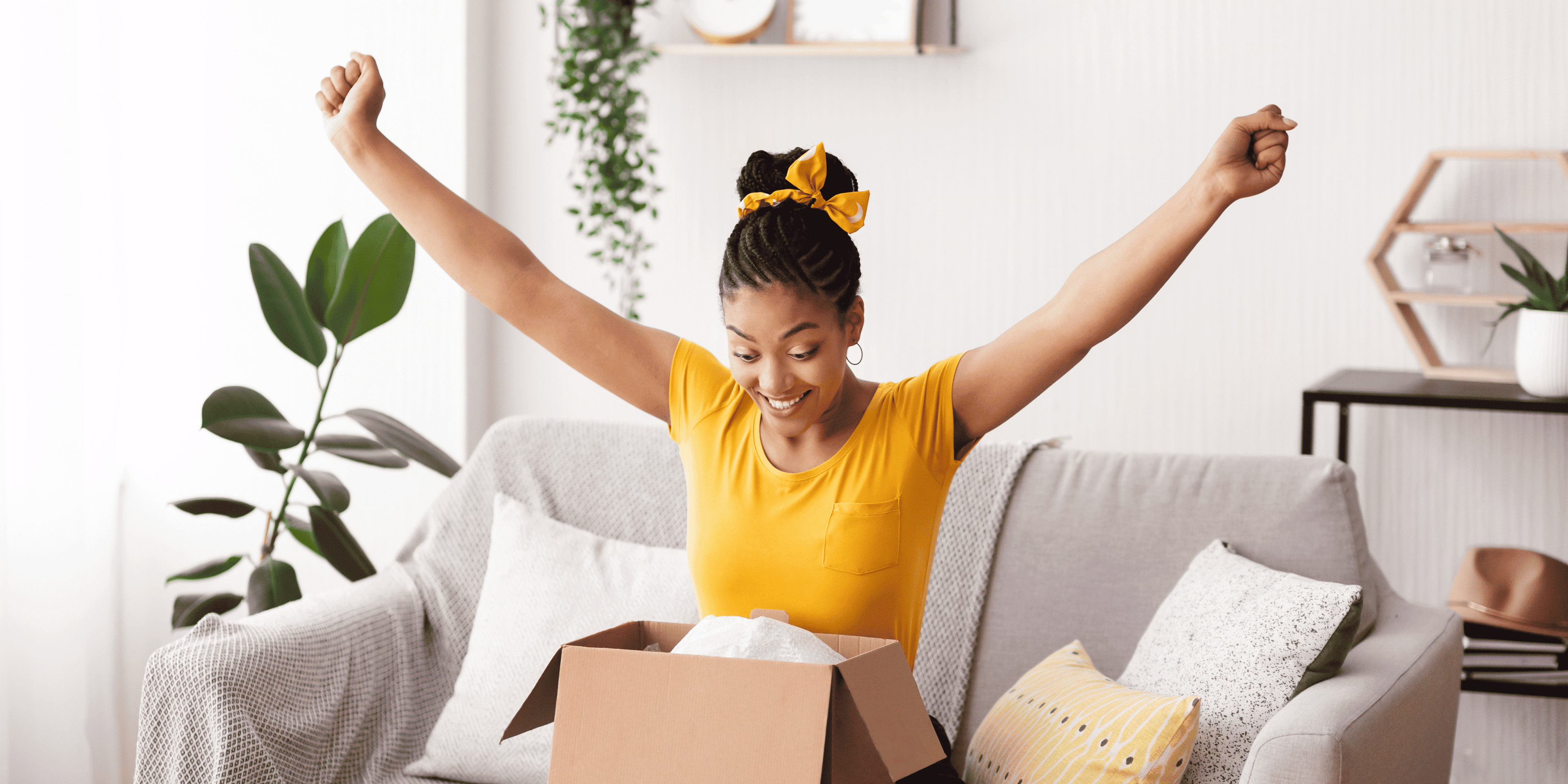 Woman in yellow shirt opening a box on a couch with arms raised in a living room.